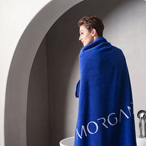 A woman wearing a blue Morgan & Tina luxurious Egyptian cotton bath towel in front of a bathtub.